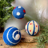 Three felted ornament balls with blue, white, and red patterns on a wooden surface with greenery.